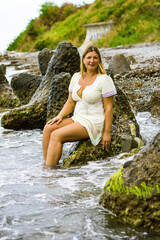 A young blonde woman in a short yellow dress sits on large stones, knee-deep in water on the seashore.