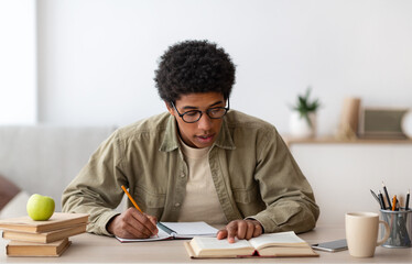 Distance learning. Black teen college student with textbook and copybook studying at home during...