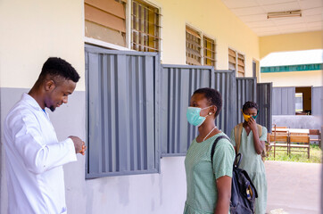 a young handsome african class teacher holding as thermometer to scan the temperature of his student before entering the classroom