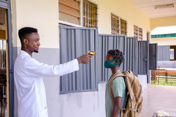a young handsome african class teacher holding as thermometer to scan the temperature of his student before entering the classroom