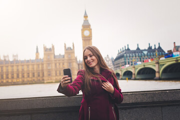Obraz premium Young London traveller woman takes selfie pictures with her phone at Westminster with view to the Big Ben Tower in London, UK