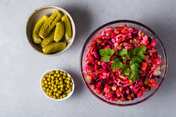 Vinigret in a glass salad bowl with a parsley leaf, pickled cucumbers in a round plate, canned peas on a gray background