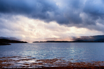 Fototapeta na wymiar Rays of sunlight break through brooding storm clouds to illuminate the loch shore lighting up the middle of the picture with gold light the blue of the storm clouds and the lake water above and below