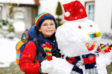 Little school kid boy in colorful clothes, with glasses and backpack having fun with snowman after elementary school end