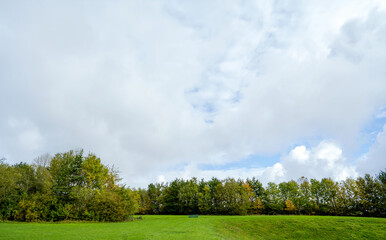 Beautiful natural landscape meadow at public park with could and blue sky in spring or summer. Fresh air panorama view of grass field with metal bench on green forest trees environment
