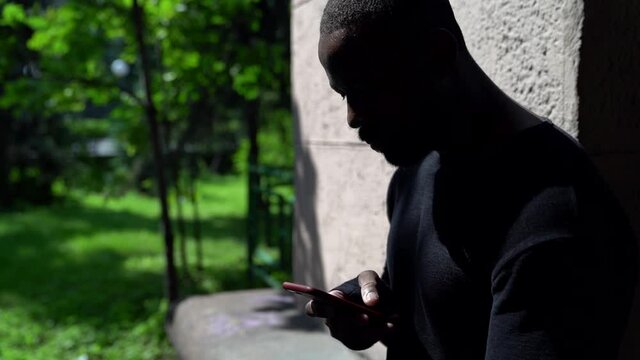 portrait of an African in dark clothing. he is sitting on a stone bench in a city Park with a red phone in his hands and typing. the camera is moving