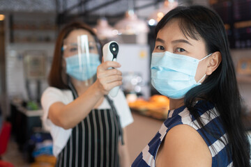 Asian waitress woman wearing face masks and holding an infrared forehead thermometer to check body...