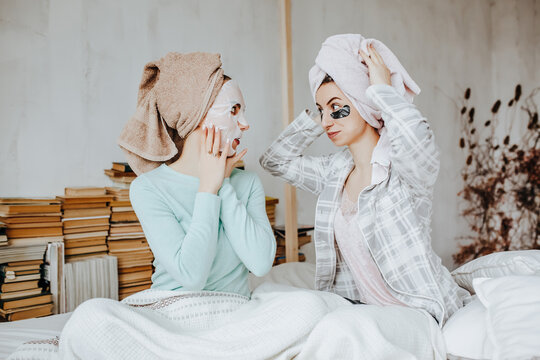 Two girls make masks and patches for the beauty of face and hair. Women take care of youthful skin. Girlfriends laugh, make faces and do beauty treatments at home with towels on their heads.
