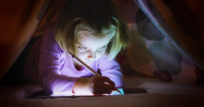 Child blonde caucasian girl digital coloring with tablet app under bed blanket at night. Modern technology childhood kid use at home. 4k video