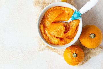 Homemade pumpkin puree in bowl with baby spoon and fresh pumpkins on light concrete background. The concept of baby food.