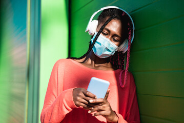 Young black woman listening music playlist with mobile phone while wearing face mask for...