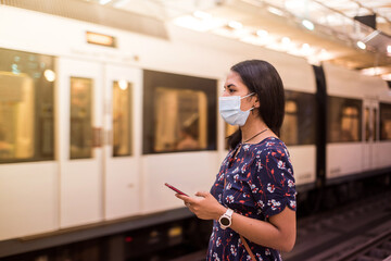 Young latin woman wearing face mask and using the smart phone on the subway station during coronavirus pandemic.