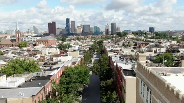 Cinematic aerial pullback reveals expanse of Baltimore skyline from Federal Hill neighborhood, colonial historic houses line street during summer day