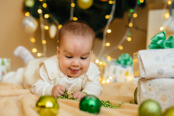 Fototapeta na wymiar Cute smiling baby is lying under a festive Christmas tree and playing with gifts. Christmas and New Year celebrations