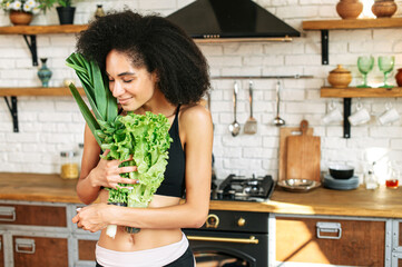 A natural beauty athletic african american woman with an armful of fresh vegetables and greenery in the kitchen at home sniffs it with eyes closed. Healthy lifestyle concept