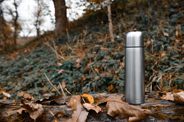 Fototapeta premium Metal vacuum thermos in the forest, copy space. A camping thermos stands on a tree outside. Hiking, active tourism, picnic, hot drink concept