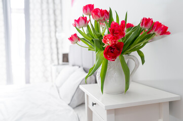 Bedroom in soft light colors..White vase with red tulips in light cozy bedroom interior. White wall, bed with white linen,