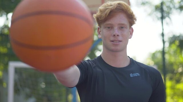 Confident Caucasian basketball player stretching ball at camera and smiling. Portrait of young cheerful man posing on sunny outdoor court on summer day. Sport and workout concept.