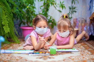 Two girls in protective medical masks paint with paints lying on the floor at home or in kindergarten