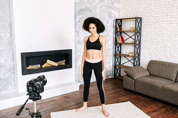 A female fitness coach records fitness video classes at home. An african american girl in sportswear is doing stretching exercises in front of camera. Sports vlogging concept, online training