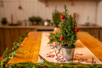 A small Christmas tree with red balls in a metal bucket stands on a wooden table in the decorated kitchen