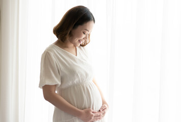 Asian pregnant woman wearing white dress and standing beside window