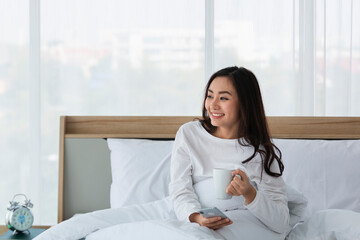 Young woman in white relaxing and drinking cup of hot coffee or tea using smartphone on bed in the bedroom. Happy girl using a mobile phone on bed. Beautiful woman using smartphone in the morning.
