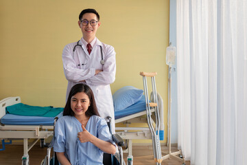 Fototapeta premium Healthcare and Treatment Concept. Young Asian female patient smiley face lifts hand up fighting with illness on wheelchair. Doctor and Patient showing fighting spirits over illness in hospital room.
