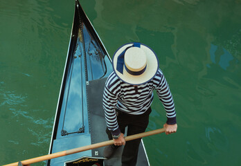 Gondolier from above. Venice iconic view. Man rowing and wearing a canotier hat and a stripes t...