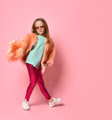 girl in stylish attire and glasses posing leaning forward on one leg and indicating at camera. Full length shot isolated on pink, copy space
