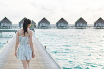 Beautiful traveler woman in a beach dress walks over a wooden pier on a tropical island. Young woman in white clothes walking on a wooden pier. Romantic honeymoon getaway in overwater bungalows villa.