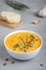 Homemade organic creamy pumpkin soup made of pureed vegetables decorated with seeds and fresh thyme served in white bowl with bread slices and garlic on concrete background. Vertical orientation