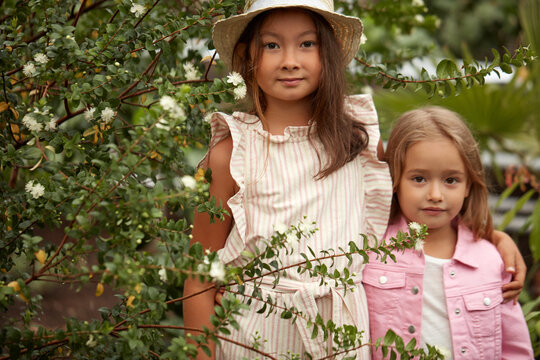 two diverse kid girls among flowers and trees, posing, sisters in casual wear have rest in the garden