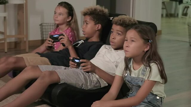 Four pretty multi racial kids play to play station seated together at home. Alpha generation offspring addicted with virtual games problem concept