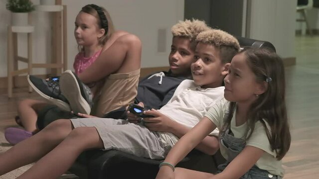 Four cute multi ethnic kids play to play station seated together at home. Alpha generation offspring addicted with virtual games problem concept