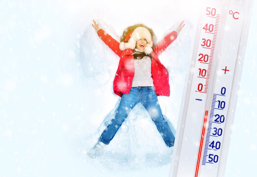 Real winter concept with a girl making snow angel in a snow and big thermometer. Еhermometer shows frosty temperature. Christmas holidays and entertainment background. Copy space.
