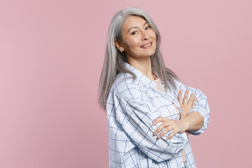 Side view of smiling beautiful gray-haired asian woman wearing basic white checkered shirt standing holding hands crossed looking camera isolated on pastel pink colour background studio portrait.