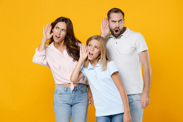 Shocked curious young parents mom dad with child kid daughter teen girl in basic t-shirts try to hear you overhear listening intently isolated on yellow background studio portrait. Family day concept.