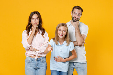 Confused young parents mom dad with laughing child kid daughter teen girl in t-shirts put hand prop up on chin pointing index finger on camera isolated on yellow background studio. Family day concept.