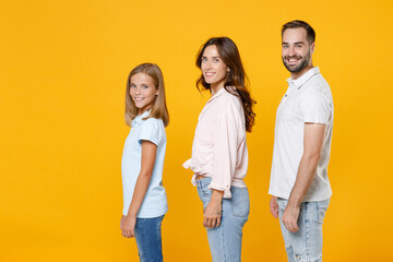Fototapeta premium Side view smiling young happy parents mom dad with child kid daughter teen girl in basic t-shirts looking camera isolated on yellow background studio portrait. Family day parenthood childhood concept.