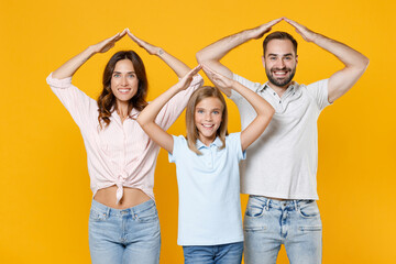 Obraz premium Smiling funny young parents mom dad with child kid daughter teen girl in basic t-shirts holding hands above head like roof of house isolated on yellow background studio portrait. Family day concept.