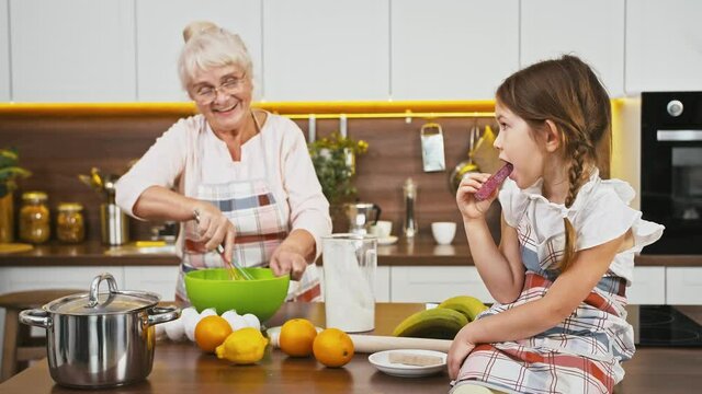Granny putting flour and egg into bowl mixing it by whisk, talking to little granddaughter who sitting on table eating candy at modern kitchen