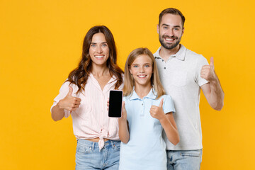 Obraz premium Smiling funny young parents mom dad with child kid daughter teen girl in basic t-shirts hold mobile phone with blank empty screen showing thumbs up isolated on yellow background. Family day concept.