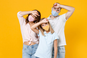 Sick tired young parents mom dad with child kid daughter teen girl in t-shirts face mask to safe from coronavirus virus covid-19 put hands on heads isolated on yellow background. Family day concept.