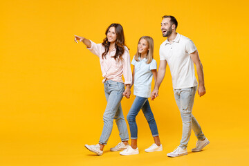 Full length portrait of smiling funny young parents mom dad with child kid daughter teen girl in t-shirts holding hands pointing index finger aside isolated on yellow background. Family day concept.