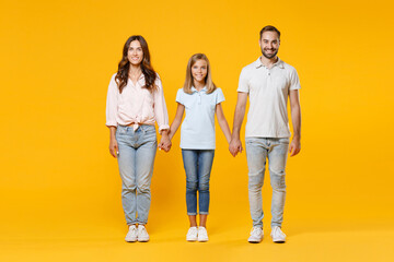 Fototapeta premium Full length portrait of smiling young parents mom dad with child kid daughter teen girl in t-shirts holding hands looking camera isolated on yellow background. Family day parenthood childhood concept.