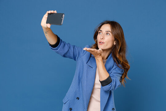 Pretty charming beautiful young brunette woman 20s wearing basic jacket doing selfie shot on mobile phone blowing sending air kiss isolated on bright blue colour wall background, studio portrait.