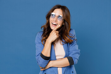 Cheerful funny excited pensive young brunette woman 20s wearing basic jacket eyeglasses standing put hand prop up on chin looking aside up isolated on bright blue colour background studio portrait.