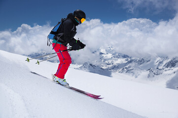 Fototapeta na wymiar woman skier athlete makes a jump in flight on a snowy slope against the backdrop of a blue sky of mountains and clouds. Freeride and extreme skiing for women