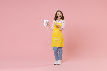 Full length portrait of excited young woman housewife wearing yellow apron pointing index finger on mixer while doing housework isolated on pastel pink colour background studio. Housekeeping concept.
